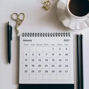Flat lay of a 2021 desk calendar, coffee cup, and black stationery items on a white surface.